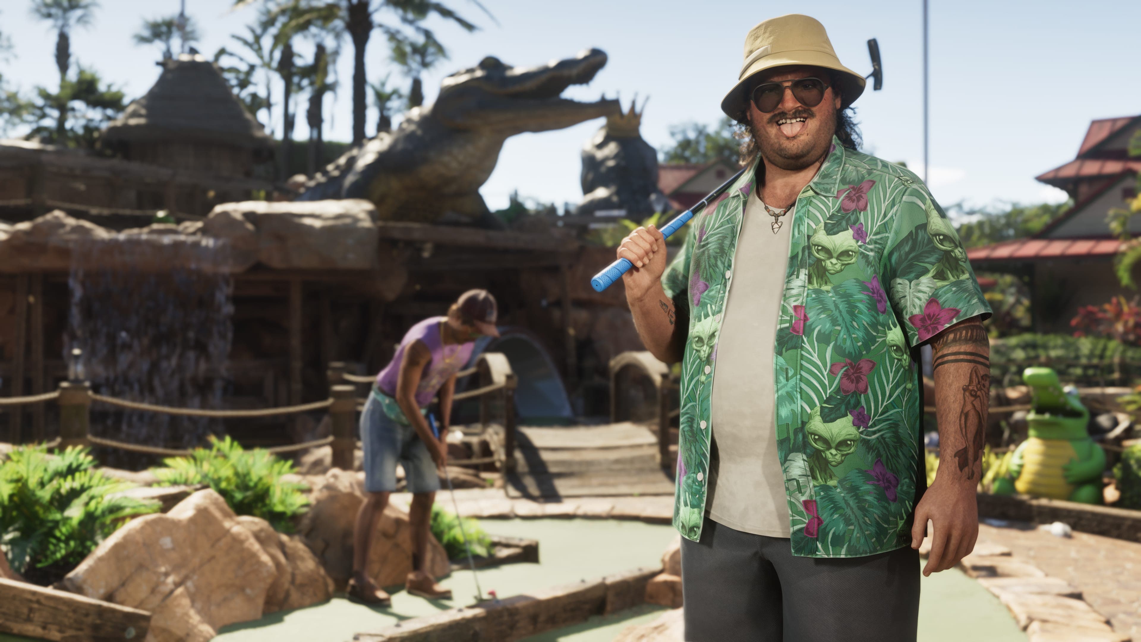 Cal Hampton in a bucket hat, sunglasses and a green shirt printed with aliens rests a club on his shoulder at a mini golf course decorated with fake alligators while another man putts