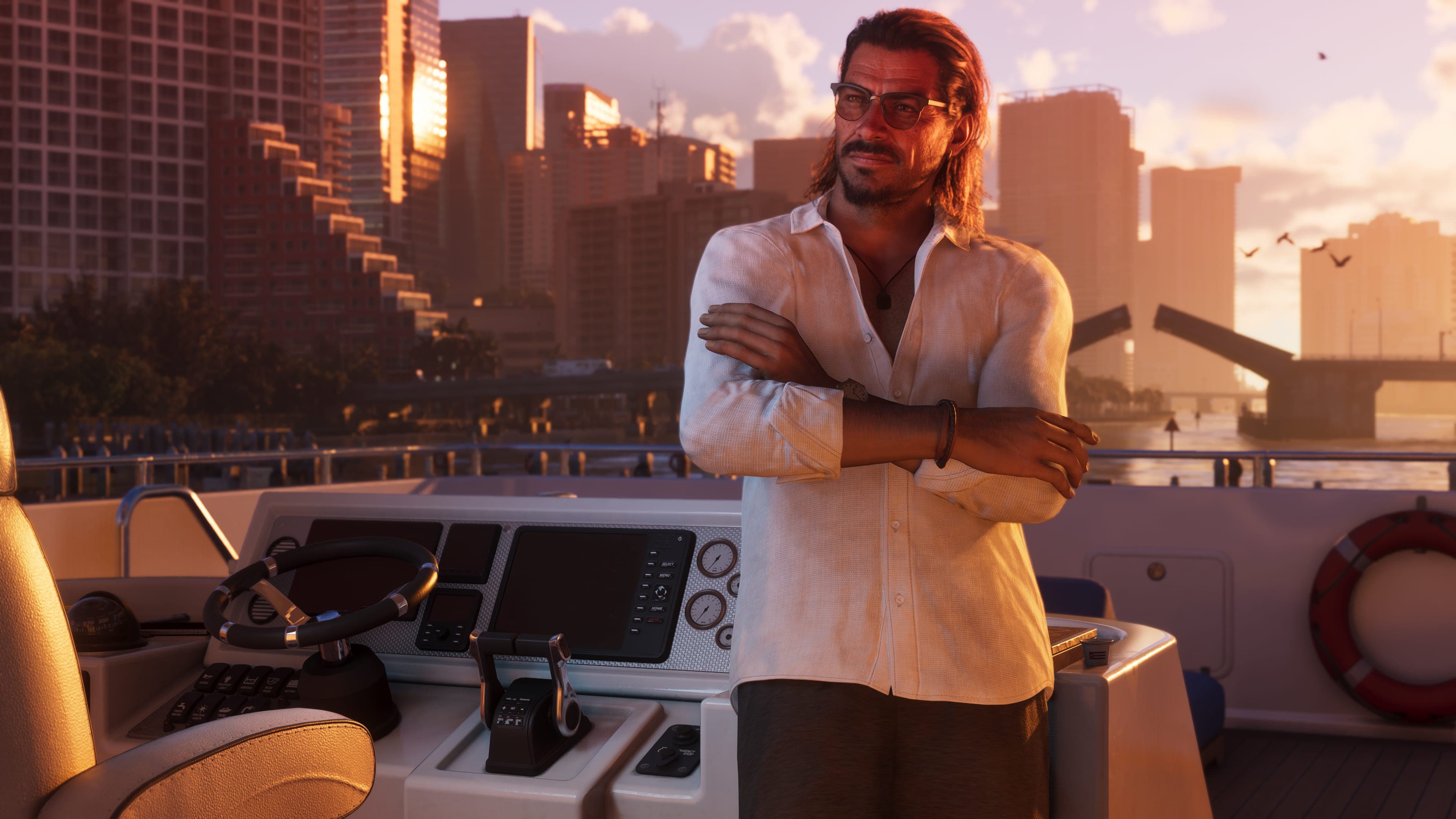 Raul Bautista in sunglasses and an open white shirt crosses his arms at the helm of a boat, with a city skyline at sunset behind him